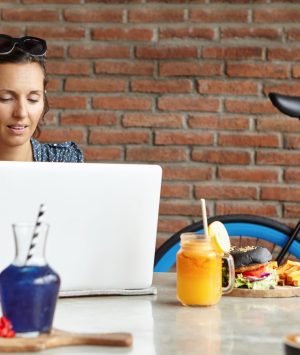 Beautiful female wearing sunglasses on her head, browsing internet, checking her newsfeed via social networks and messaging online, using free wi-fi at modern coffee shop, having lunch alone